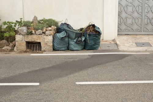 The image shows three large dark green garbage bags filled with rubbish, placed on the pavement adjacent to a white wall. The bags appear to be made from durable plastic, with some contents slightly bulging and visible at the openings, indicating they are full. To the left of the bags, there is a small, rocky garden bed with a few green plants growing amidst stones and soil. Behind the garden area, there is a small rectangular vent embedded in the wall below the plants. To the right of the rubbish bags, part of a decorative grey metal gate is visible, set into the white wall, adjacent to a paved sidewalk. The scene is lit by natural daylight, with shadows cast on the pavement, suggesting clear weather. The setting appears to be an exterior urban or suburban street, where Garden Clearance Brompton services might be engaged for rubbish removal, illustrating typical household or garden waste awaiting collection.