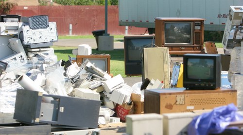 A large collection of discarded electronic waste, including old CRT televisions with wooden casings, smaller box-style monitors, and various outdated computer components, all piled outdoors on the pavement. The pile contains a mix of beige, brown, and black devices, some stacked atop one another and others leaning or scattered around. In the background, a red brick wall and a green grassy area are visible, indicating the waste is situated in an open outdoor space, possibly near a residential or industrial area. The image captures the cluttered nature of electronic rubbish awaiting collection or disposal, with no visible vehicles or tools directly associated with rubbish removal. This scene aligns with the services offered by Garden Clearance Brompton, especially in managing rubbish including electronic waste for clients within the Brompton area, elsewhere in the vicinity.