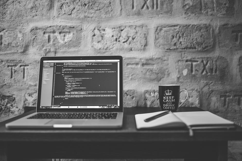 A man with glasses and a beard is seated at a wooden desk, working on a computer with dual monitors. The left monitor displays a file management interface with a dark theme, while the right monitor sh