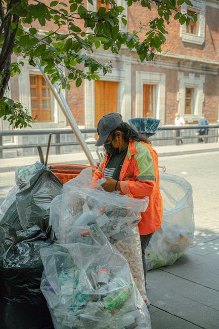 A middle-aged man with dark hair, wearing a black T-shirt with graphic white text on the front, is seen bending over to dispose of a white plastic bag filled with rubbish into a stainless steel litter