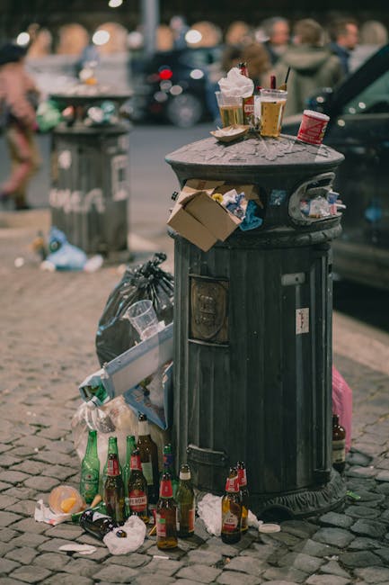 A pile of mixed waste materials located at the base of a brick and concrete wall, including cardboard boxes, some of which are flattened and partially open, and plastic bags. The cards are in various 